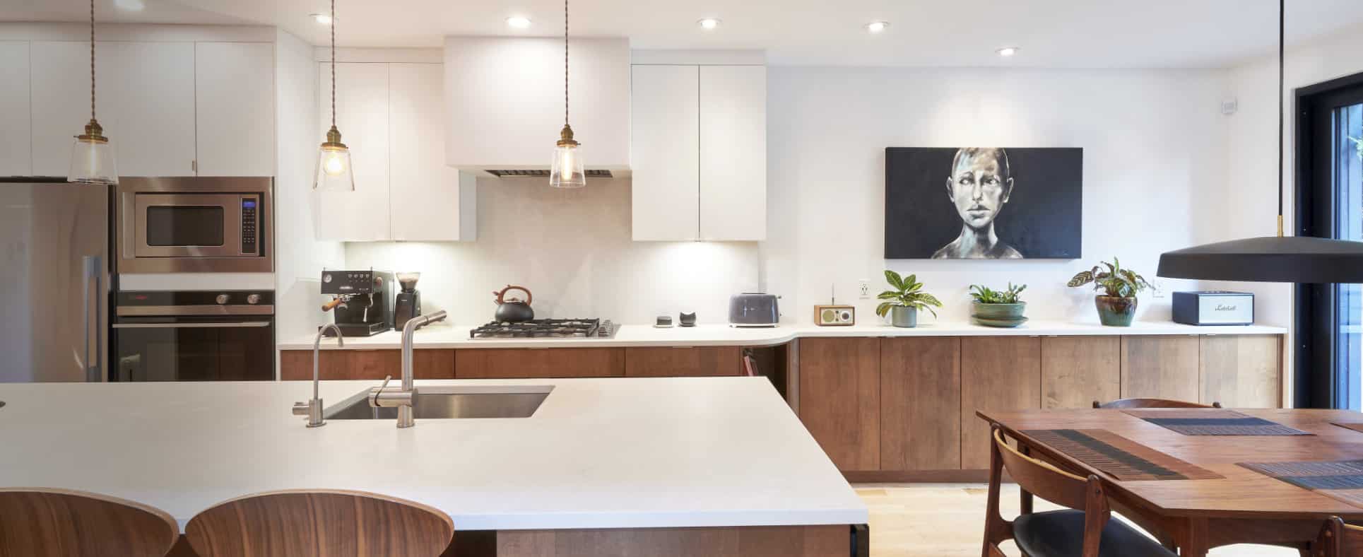 Cuisine moderne avec un grand îlot, des comptoirs blancs et des armoires en bois. Des électroménagers en acier inoxydable sont intégrés à la conception. Des luminaires suspendus illuminent l'espace. Un tableau et des plantes décorent le comptoir.