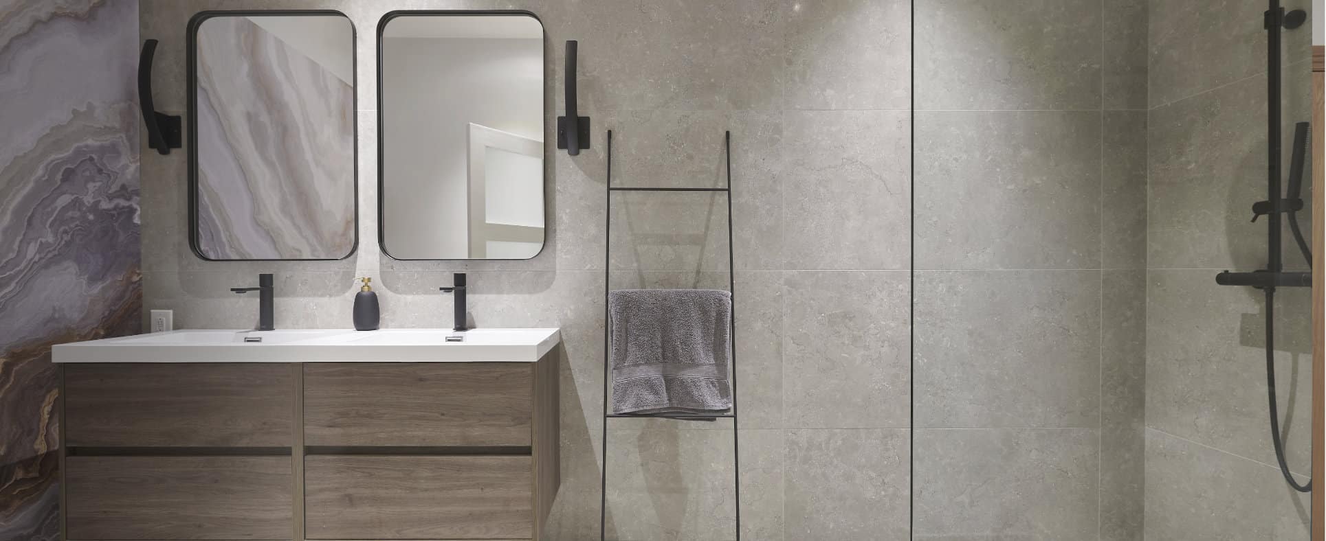 Salle de bain moderne avec une double vasque dotée de deux miroirs rectangulaires et de robinets noirs. La vanité est dotée de tiroirs en bois. Un élégant porte-serviettes accueille une serviette grise. La pièce est dotée d'un mur en carrelage de pierre grise et d'une cabine de douche en verre.