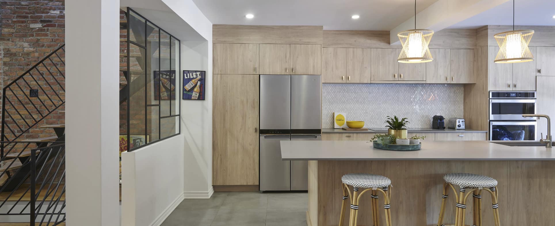 Cuisine moderne avec des meubles en bois clair, des appareils électroménagers en acier inoxydable et un grand îlot avec deux tabourets à motifs. Des luminaires suspendus sont suspendus au-dessus de l'îlot et on aperçoit un escalier avec un mur de briques en arrière-plan.