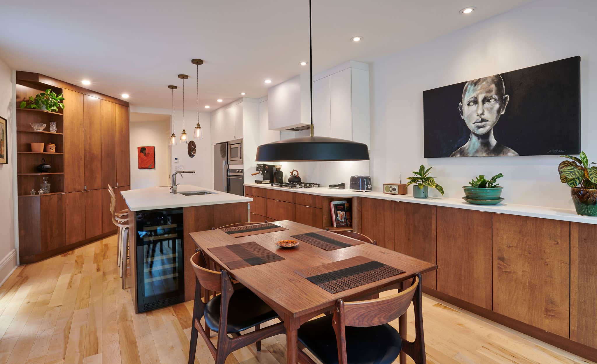 Une cuisine moderne avec des meubles en bois et un îlot avec un évier et des tabourets de bar. Une table à manger avec des chaises est au premier plan. Des luminaires suspendus sont suspendus au plafond. Il y a des œuvres d'art sur les murs et des plantes sur les comptoirs.