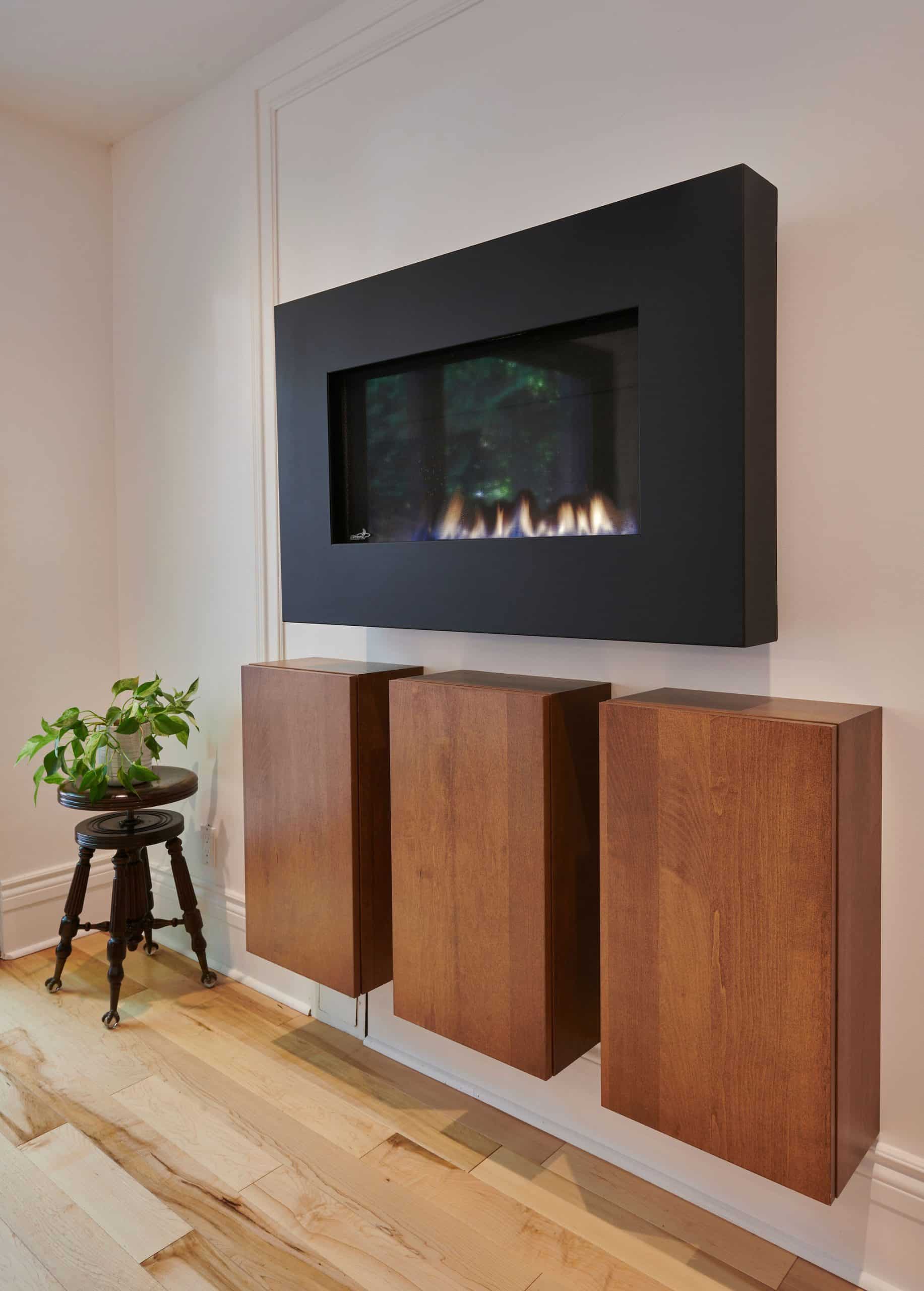 Une cheminée moderne avec un cadre noir épuré, montée au-dessus de trois armoires en bois sur un mur de couleur claire. Une petite plante verte est posée sur un tabouret en bois foncé, ajoutant une touche naturelle à la décoration intérieure minimaliste.