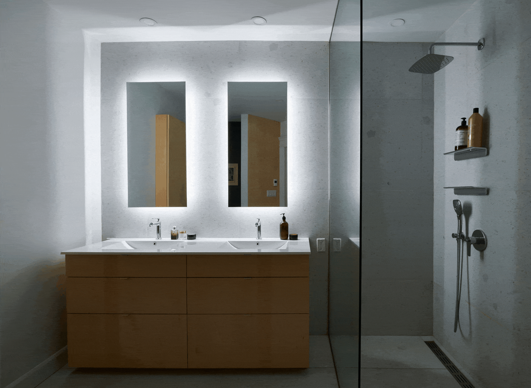 Salle de bains moderne avec meuble-lavabo double en bois et deux miroirs éclairés. Une douche vitrée avec pommeau de douche à effet pluie et étagères pour les produits de toilette se trouve à droite. L'éclairage est doux, créant une atmosphère sereine.