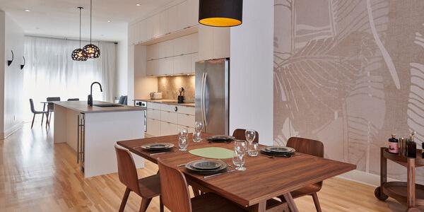 Une salle à manger et une cuisine modernes avec parquet en bois présentent une transformation étonnante. La table à manger est dressée avec des assiettes et des verres. La cuisine dispose d'armoires blanches, d'un réfrigérateur en acier inoxydable et de deux luminaires suspendus au-dessus de l'îlot. Une grande fresque murale décore élégamment un mur.