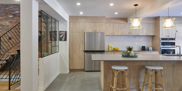 Intérieur de cuisine moderne à Mont-Royal avec armoires en bois clair, électroménagers en acier inoxydable et sol en carrelage gris. Deux tabourets de bar en osier sont installés sur un îlot spacieux. Des luminaires suspendus sont suspendus au-dessus et un escalier avec une rampe noire est visible sur la gauche, se fondant parfaitement dans le style de la rénovation.