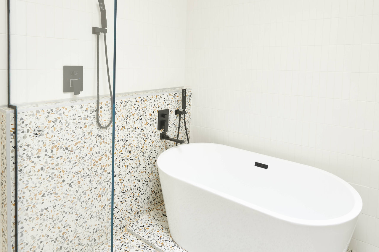 Une salle de bains moderne avec une baignoire îlot blanche à côté d'un rebord carrelé en terrazzo et une douche vitrée. La douche est dotée d'un robinet et d'un pommeau de douche noirs élégants, et les murs sont tapissés de grands carreaux blancs.