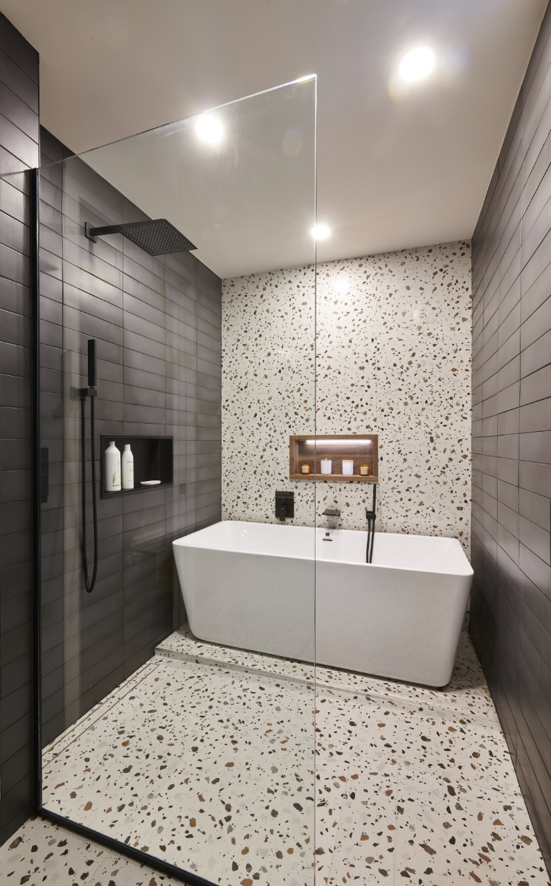 Salle de bains moderne avec sol et murs en terrazzo, dotée d'une baignoire îlot blanche et d'une élégante pomme de douche à effet pluie noire. L'espace est éclairé par des plafonniers encastrés, mettant en valeur son design épuré et minimaliste.