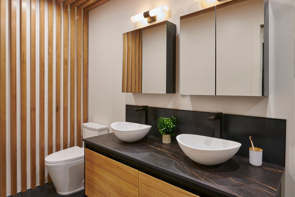 Salle de bains moderne avec deux vasques blanches sur un plan de travail en marbre noir, des armoires en bois et un mur d'accent en lattes de bois. Au-dessus des lavabos se trouvent des armoires à miroir et des luminaires muraux. Une petite plante et un porte-brosse à dents sont posés sur le comptoir.