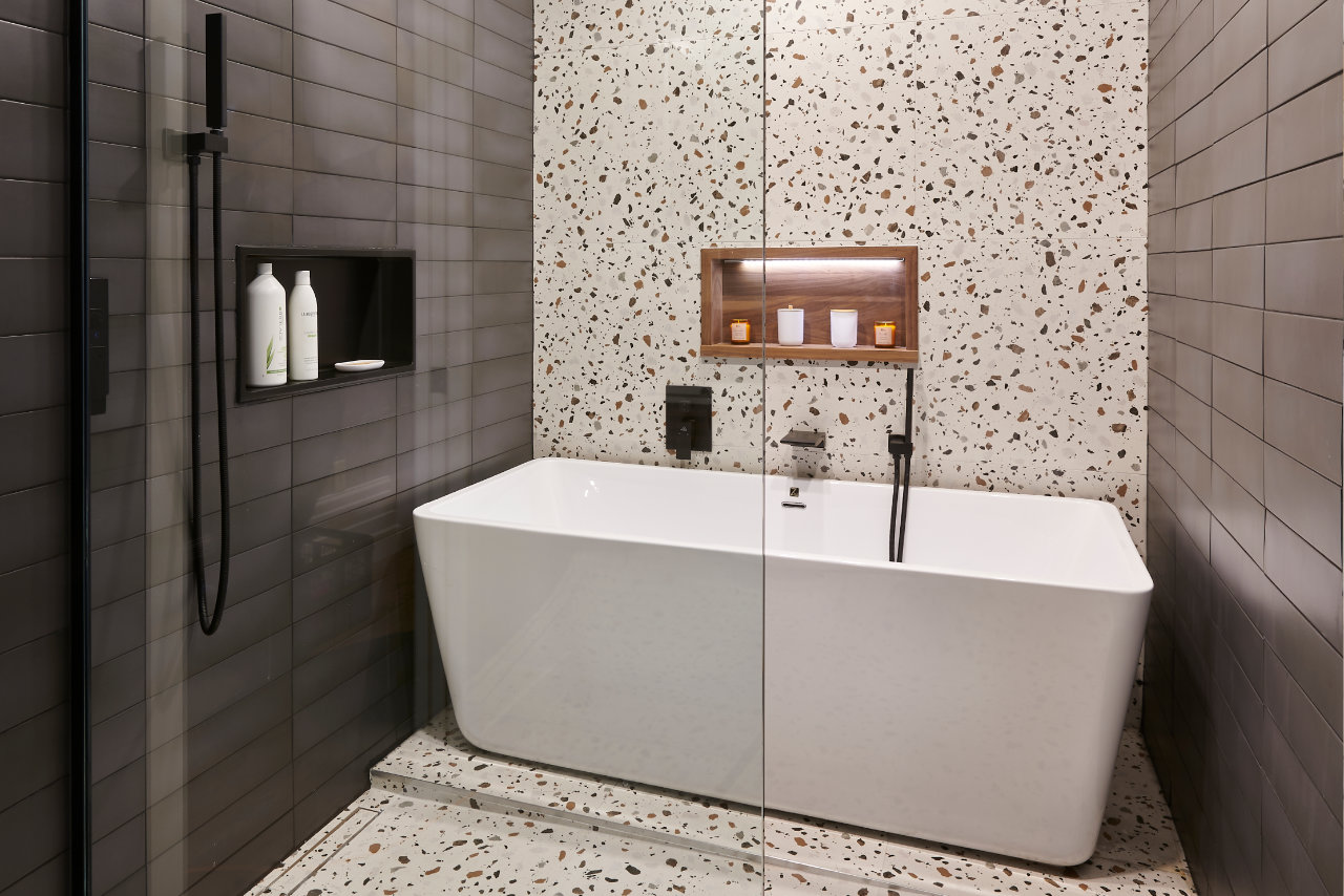 Salle de bains moderne avec sol et murs en terrazzo, dotée d'une baignoire îlot blanche et d'une élégante pomme de douche à effet pluie noire. L'espace est éclairé par des plafonniers encastrés, mettant en valeur son design épuré et minimaliste.