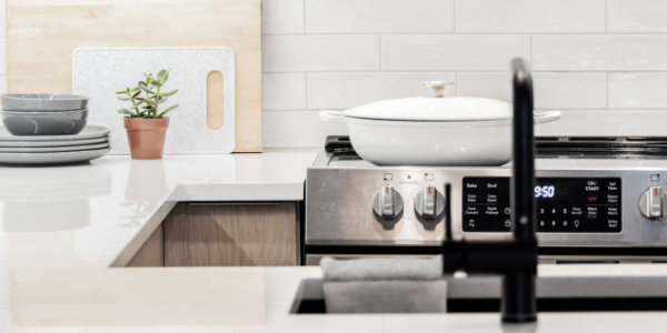 Cuisine moderne avec une cuisinière en acier inoxydable. Dosseret en carreaux de métro blancs et armoires en bois clair. Une plante en pot, des assiettes et des planches à découper sont posées sur le comptoir. Un évier et un robinet noirs se trouvent au premier plan.