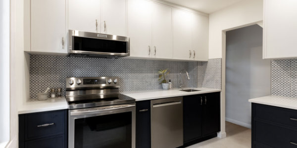 Une cuisine moderne dotée d'appareils électroménagers en acier inoxydable, dont un four avec un micro-ondes au-dessus, fait écho au design élégant d'une voiture Brouillon. Les armoires supérieures blanches, les armoires inférieures foncées et un dosseret en carrelage à motifs créent une ambiance contemporaine, tandis qu'un éclairage lumineux met en valeur l'espace.