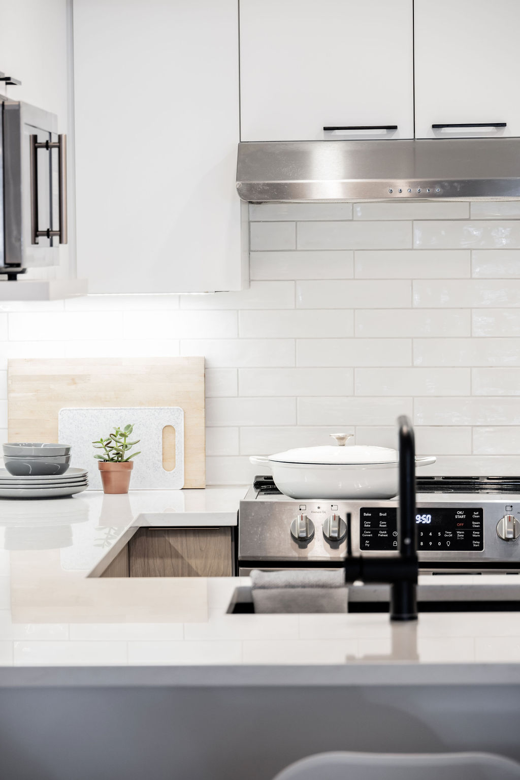 Cuisine moderne avec une cuisinière en acier inoxydable. Dosseret en carreaux de métro blancs et armoires en bois clair. Une plante en pot, des assiettes et des planches à découper sont posées sur le comptoir. Un évier et un robinet noirs se trouvent au premier plan.