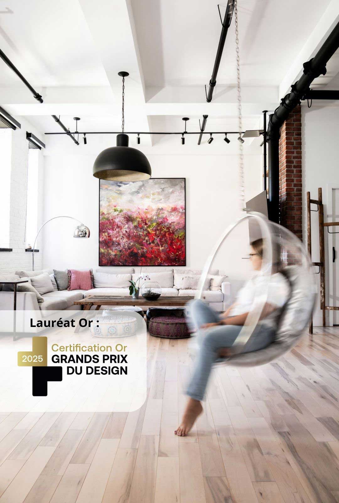 Un salon moderne avec un fauteuil suspendu en forme de bulle en mouvement, occupé par une personne. L'espace comprend un parquet en bois clair, un canapé sectionnel gris, des coussins colorés, des poufs ronds, un grand tableau abstrait et un éclairage et des tuyaux de style industriel.