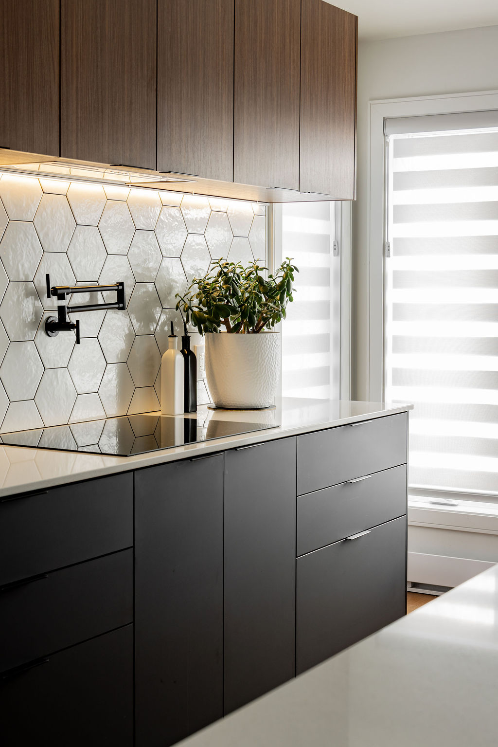 Plaque d'induction avec remplisseur de casserole mural noir mat, dosseret hexagonal blanc brillant et éclairage DEL sous caisson en bois, conçu par un designer d'intérieur à Montréal.