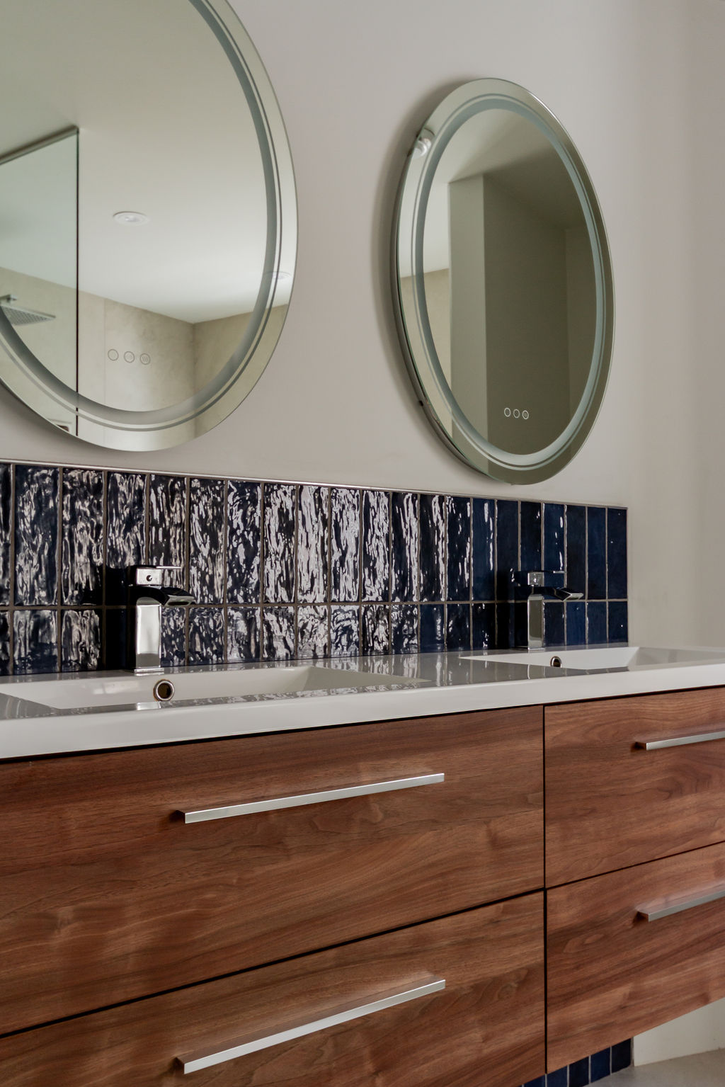 Vanité double en bois de noyer suspendue avec dosseret en tuiles bleu marin texturées et miroirs ovales lumineux LED, conçue par un designer d'intérieur à Montréal.