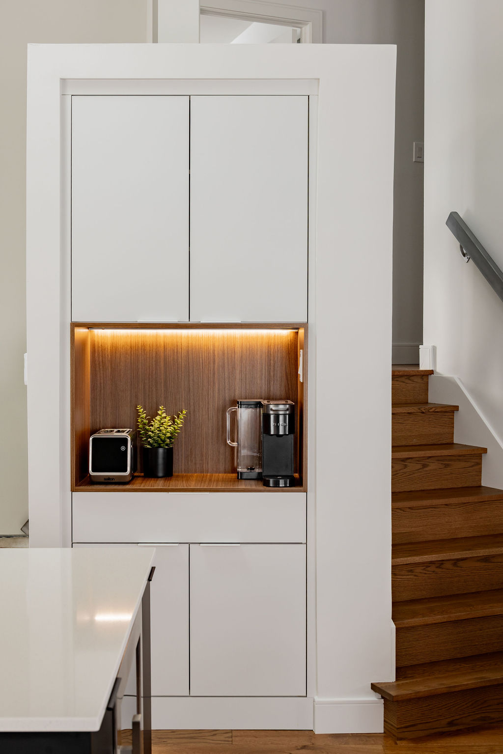 Coin café encastré sur mesure dans un mur par un designer d'intérieur à Montréal, avec armoires blanches, niche en bois chaleureux rétroéclairée à DEL et escalier en bois assorti.