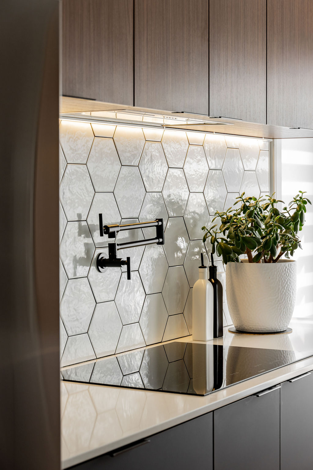Plaque d'induction avec remplisseur de casserole mural noir mat, dosseret hexagonal blanc brillant et éclairage DEL sous caisson en bois, conçu par un designer d'intérieur à Montréal.
