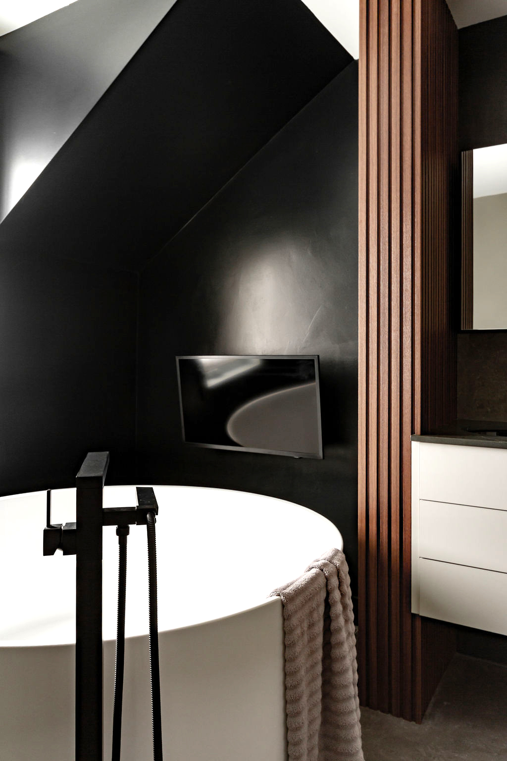 Salle de bain moderne haut de gamme à Montréal par un designer d'intérieur, avec une baignoire cylindrique autoportante blanche, un robinet autoportant noir mat, et une fenêtre de coin sous un plafond incliné.